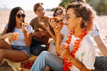 Group of young friends eating pizza, toasting with beerus and have fun on the beach. Happy friends resting together sitting near the sea. Fast food concept. Summer holidays, vacation, relax.