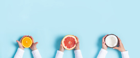 Canvas Print - childrens hands hold orange, grapefruit and coconut on a blue background. Top view, flat lay. Banner