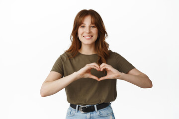 Wall Mural - Cute young woman smiling, show heart I love you sign, like or care for someone, standing in t-shirt against white background