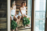 Young women friends in white towel sitting on wooden shelf in Finnish sauna Russian bath with rowan brooms in spa hotel in front of the panoramic window, treat yourself