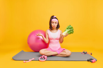 Puzzled fit Asian woman sits in lotus pose on fitness mat holds green salad surrounded by sport equipment keeps to healthy diet has regular exercises stays at home during quarantine yellow background