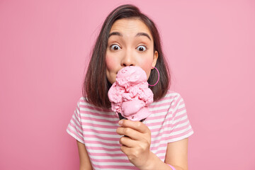 Photo of surprised Asian woman licks delicious ice cream has sweet tooth eats high calorie food dressed in casual striped t shirt during summer isolated on pink background. Treat yourself with dessert