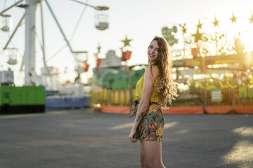 Wall Mural - Positive young female in summer dress standing over Ferris wheel in amusement park and having fun