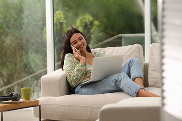 Wall Mural - Young woman talking on phone while working with laptop at home