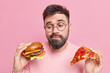 © WHstudio Leushin N - Handsome bearded adult European man feels hesitant whether to eat hamburger or pizza prefers eating junk food wears round spectacles and jumper isolated over pink background. Cheat meal concept