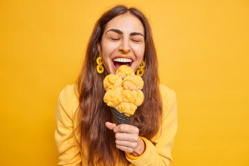 Wall Mural - Overjoyed brunette woman eats tasty ice cream on mango flavor bites appetizing snack has dirty face foolishes around keeps eyes closed isolated over vivid yellow background. Summer time concept