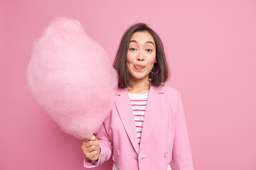 Wall Mural - Elegant businesswoman has day off licks lips starves for eating delicious sugary cotton candy wears stylish jacket poses against pink background. Cute young Asian woman with tasty sugary dessert