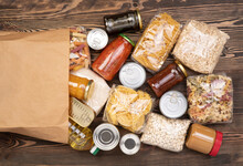 Food Donations Such As Pasta, Rice, Oil, Peanut Butter, Canned Food, Jam In A Paper Bag On Brown Wooden Table 
