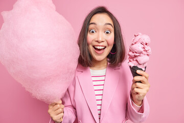 Wall Mural - Studio shot of happy surprised young Asian woman with dark hair stares impressed at camera holds cotton candy and tasty appetizing ice cream dressed formally isolated over pink studio background.