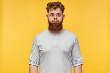 young bearded guy, with stylish hairstyle, starring into camera with widely opened eyes, puff out. Isolated over yellow background.