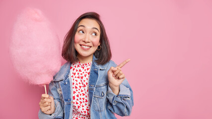 Pretty thoughtful Asian woman with cheerful expression smiles pleasantly indicates away on copy space shows direction holds delicious cotton candy wears denim jacket isolated over pink background
