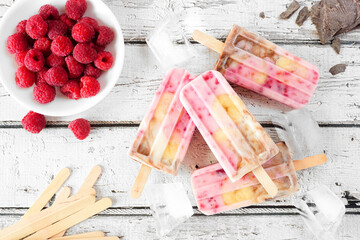 Homemade raspberry, chocolate, banana yogurt ice pops. Above view table scene on a rustic white wood background.