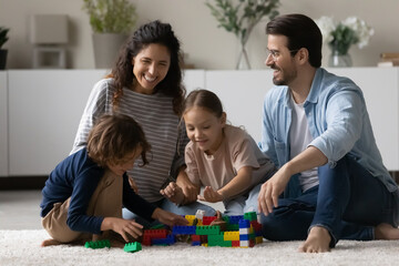 Wall Mural - Overjoyed multiethnic parents sit on floor play toys with little biracial kids together. Happy multiracial young family with small ethnic children have fun engaged in playful game activity at home.