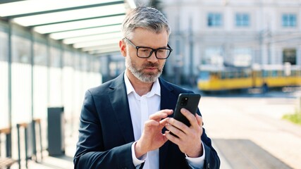 Close up portrait of happy Caucasian middle-aged stylish male traveller texting on smartphone browsing online standing in town on train station, looking at camera and smiling, traveling, business trip