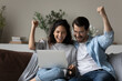 Excited multiethnic couple feel euphoric win online lottery win or victory on laptop gadget. Overjoyed multiracial man and woman relax on couch at home triumph read good news on computer.