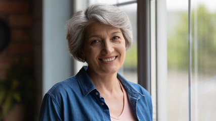 Elderly 60s woman with short haircut smile look at camera enjoy peaceful midlife retired life standing near window posing alone indoors. Head shot portrait optimistic happy senior latin female concept
