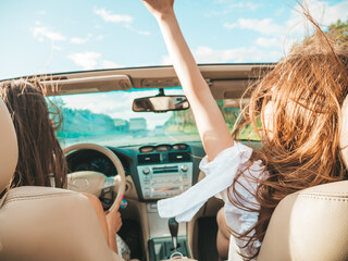 Portrait of two young beautiful and smiling hipster female in convertible car. Sexy carefree women driving cabriolet. Positive models riding and having fun in sunglasses outdoors. Enjoying summer days