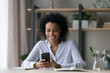 © fizkes - Smiling young African American woman sit at desk at home office text message on smartphone online. Happy millennial mixed race female distracted from work studying use modern cellphone gadget.