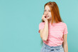Little puzzled confused redhead kid girl 12-13 years old wearing pink striped t-shirt look aside biting nails fingers isolated on pastel blue background studio. Children lifestyle childhood concept.