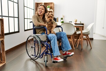 Canvas Print - Young hispanic girl sitting on wheelchair at home puffing cheeks with funny face. mouth inflated with air, crazy expression.