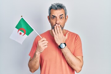 Poster - Handsome middle age man with grey hair holding algeria flag covering mouth with hand, shocked and afraid for mistake. surprised expression