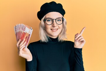 Sticker - Young blonde girl holding norwegian krone banknotes smiling happy pointing with hand and finger to the side