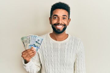 Wall Mural - Handsome hispanic man with beard holding dollars looking positive and happy standing and smiling with a confident smile showing teeth