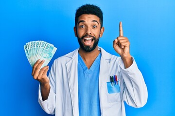 Canvas Print - Handsome hispanic man with beard wearing doctor uniform holding 100 brazilian reals smiling with an idea or question pointing finger with happy face, number one