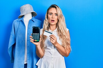 Canvas Print - Beautiful young blonde shop assistance woman holding dataphone and credit card in shock face, looking skeptical and sarcastic, surprised with open mouth