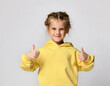 Portrait of a pretty attractive little girl in a yellow warm hoodie showing thumbs up with two hands on a white background