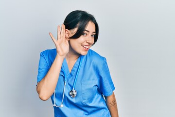 Beautiful hispanic woman wearing doctor uniform and stethoscope smiling with hand over ear listening and hearing to rumor or gossip. deafness concept.