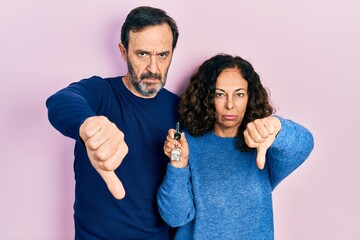 Sticker - Middle age couple of hispanic woman and man holding keys of new home looking unhappy and angry showing rejection and negative with thumbs down gesture. bad expression.