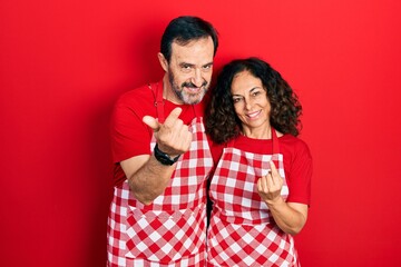 Wall Mural - Middle age couple of hispanic woman and man wearing cook apron beckoning come here gesture with hand inviting welcoming happy and smiling