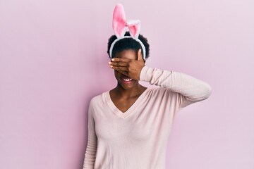 Poster - Young african american girl wearing cute easter bunny ears smiling and laughing with hand on face covering eyes for surprise. blind concept.