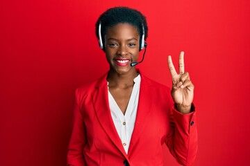 Canvas Print - Young african american girl wearing call center agent headset smiling with happy face winking at the camera doing victory sign. number two.