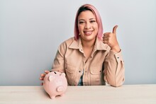 Hispanic Woman With Pink Hair Holding Piggy Bank Smiling Happy And Positive, Thumb Up Doing Excellent And Approval Sign