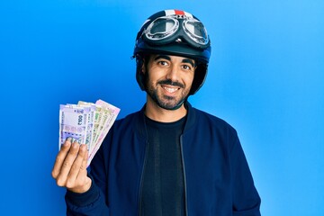 Sticker - Young hispanic man wearing motorcycle helmet holding indian rupee looking positive and happy standing and smiling with a confident smile showing teeth