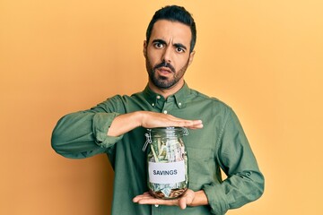 Sticker - Young hispanic man holding jar with savings in shock face, looking skeptical and sarcastic, surprised with open mouth