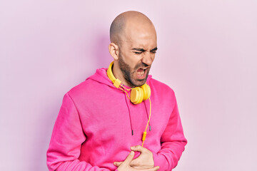 Sticker - Young bald man wearing gym clothes and using headphones with hand on stomach because nausea, painful disease feeling unwell. ache concept.