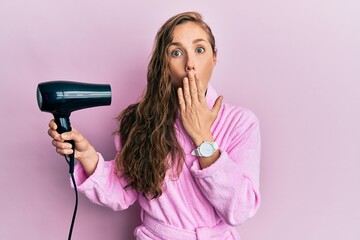 Poster - Young blonde woman wearing bathrobe using hair dryer covering mouth with hand, shocked and afraid for mistake. surprised expression