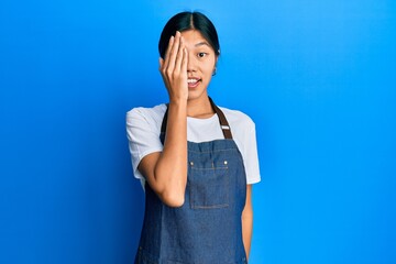 Poster - Young chinese woman wearing waiter apron covering one eye with hand, confident smile on face and surprise emotion.