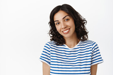 Close up portrait of attractive brunette girl with curly hair, smiling white teeth, looking cute and silly at camera, standing happy over white background