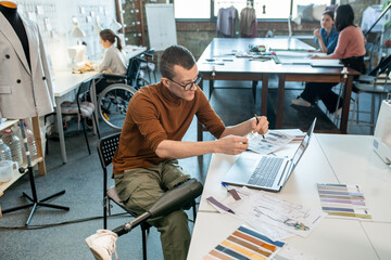 Young disable man sitting by table and working over fashion collection in large workshop