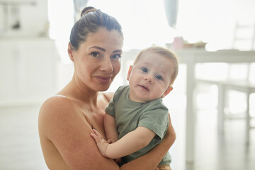 candid portrait of freckled adult mother holding baby boy and looking at camera in cozy home interio