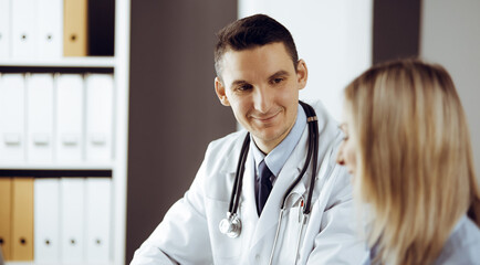 Friendly male doctor and patient woman discussing current health examination while sitting in sunny clinic. Perfect medical service in hospital