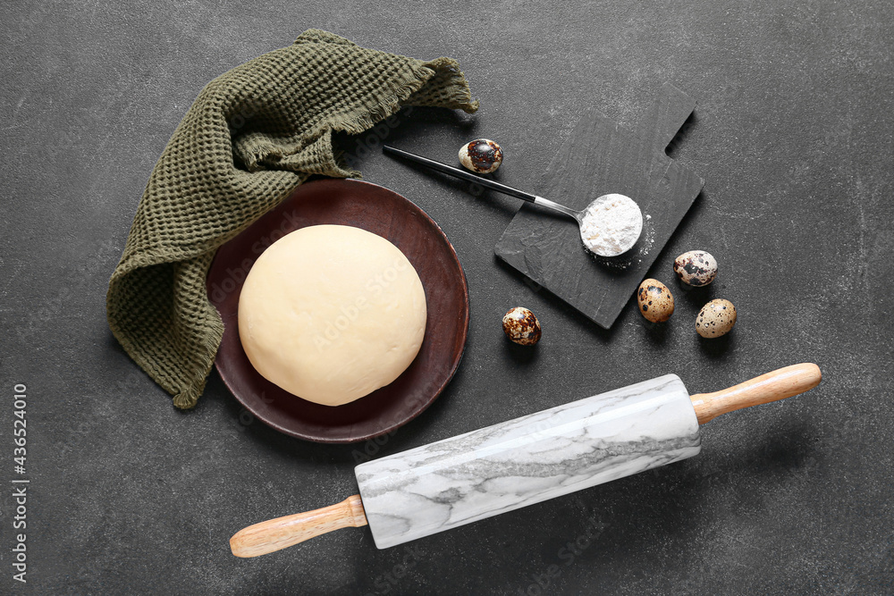 Plate with fresh dough, flour and quail eggs on dark background