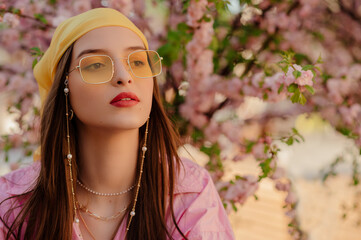 Fashionable elegant woman wearing stylish yellow bandana, trendy color glasses with chain, posing near blooming tree. Spring, summer fashion, lifestyle conception. Outdoor portrait. Copy, empty space