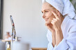 © insta_photos - Gorgeous mid age older adult 50 years old blonde woman wears bathrobe in bathroom applying nourishing antiage face skin care cream treatment, looking at mirror doing daily morning beauty routine.