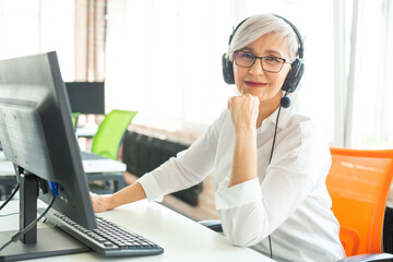 adult gray-haired woman at the age of working in the office at the computer in headphones with a microphone 