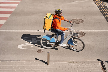 Courier man with big yellow backpack crossing road at the bicycle